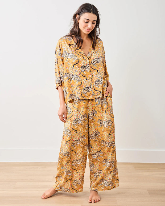 A woman in the PRINTFRESH - Bagheera Satin Wildest Dream Pajama Set in Tobacco sits barefoot on a blue velvet fringe-trimmed couch, flanked by teal geometric pillows. Blue curtains and patterned wallpaper provide a backdrop, creating a cozy ambiance that reflects the eco-friendly elegance of this PRINTFRESH pajama set.