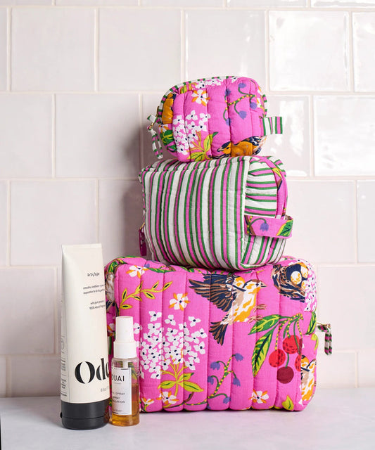A person in a white shirt holds the PRINTFRESH - BIRD & BLOSSOM THREE POUCH SET by PRINTFRESH, featuring three quilted makeup pouches with colorful floral and striped patterns, nested from smallest to largest.