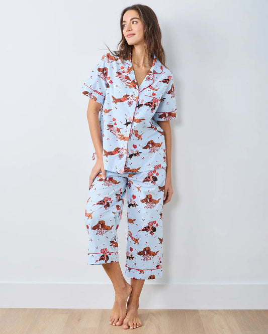 A woman with long, wavy brown hair stands barefoot against a white wall, wearing the PRINTFRESH Dachshund Date Short Sleeve Top & Cropped Pants Pajama Set in Blue. The organic cotton set from PRINTFRESH features short sleeves and cropped pants adorned with a charming dachshund print. She smiles slightly.