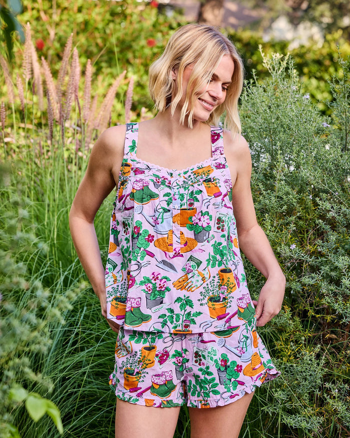 A woman with long blonde hair stands against a plain white wall, looking slightly to the side and smiling softly, wearing the PRINTFRESH - GREEN THUMB BACK TO BED PAJAMA SHORT SET by PRINTFRESH—a colorful floral sleepwear set inspired by vintage styles.
