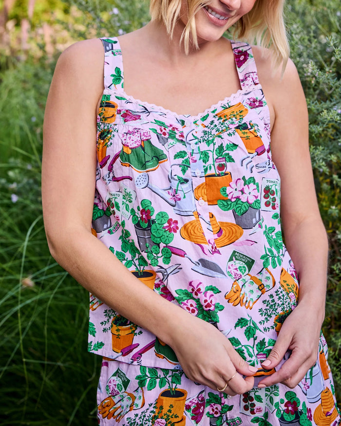 Wearing the PRINTFRESH Green Thumb Back to Bed Pajama Short Set—a vibrant, printed sleepwear ensemble from PRINTFRESH—a woman stands smiling in a lush, green garden, glancing to the side.
