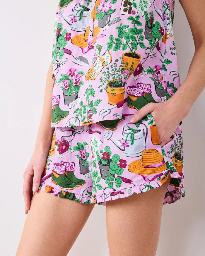 Wearing the PRINTFRESH Green Thumb Back to Bed Pajama Short Set—a vibrant, printed sleepwear ensemble from PRINTFRESH—a woman stands smiling in a lush, green garden, glancing to the side.