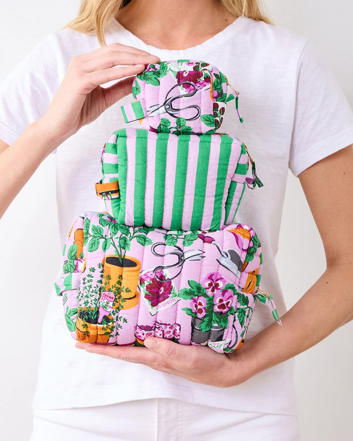 A person in a white shirt holds the PRINTFRESH - GREEN THUMB THREE POUCH SET by PRINTFRESH, featuring three quilted travel pouches with floral and striped patterns, stacked from largest to smallest, against a plain background.