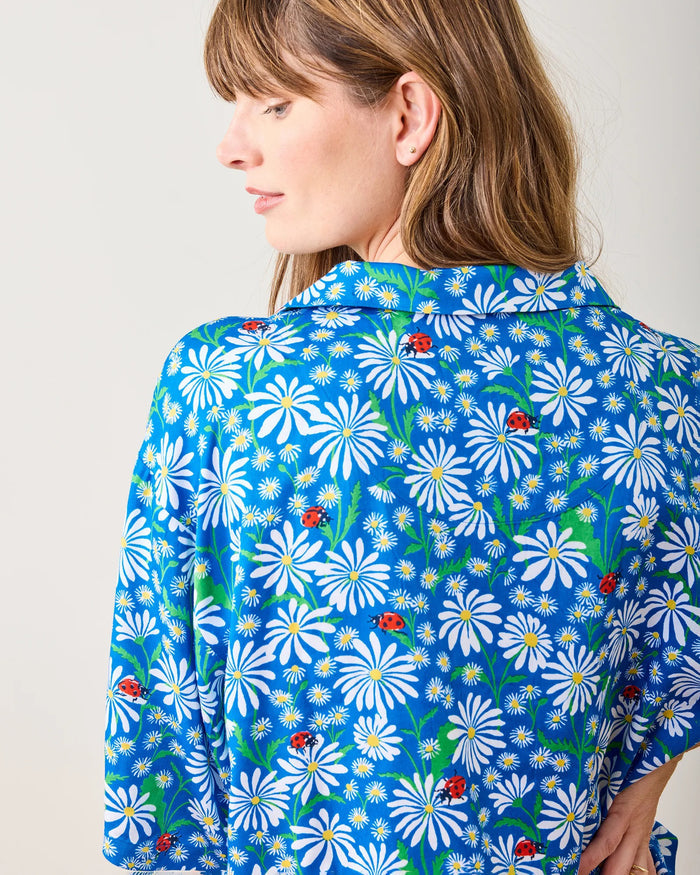 A woman stands barefoot indoors, leaning slightly against a white wall, wearing the PRINTFRESH - Lucky Daisy Satin Pajama Short Set—a blue floral matching pajamas set with shorts and a button-up top from PRINTFRESH.