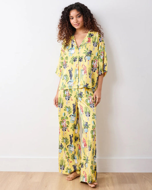 A person wears the PRINTFRESH - Macrame Mischief Satin Wildest Dream Pajama Set in Sunshine, featuring a vibrant cat and hanging plant pattern; their face is partially out of frame as they gently hold the shirt near the collar.