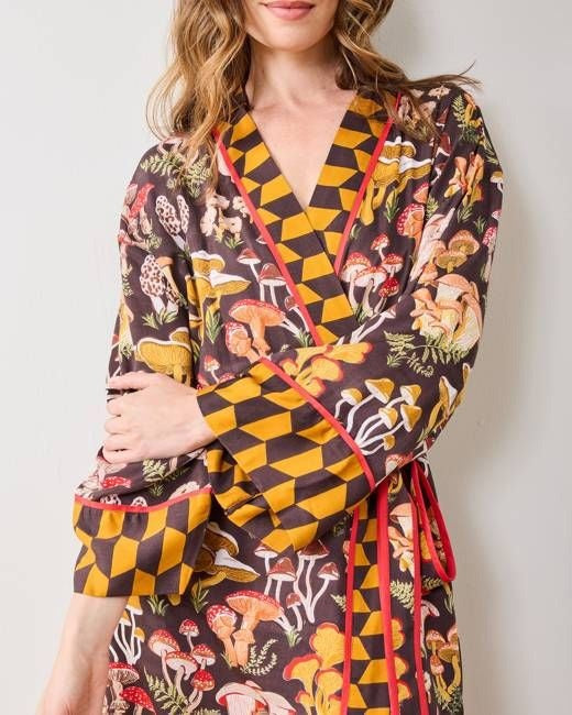 A woman is shown from the waist up wearing the PRINTFRESH - MUSHROOM MEDLEY ECO SATIN ROBE by PRINTFRESH, a kimono-style cottage-core robe featuring yellow geometric and colorful mushroom print patterns, red trim, and a tied belt. She stands against a plain wall.