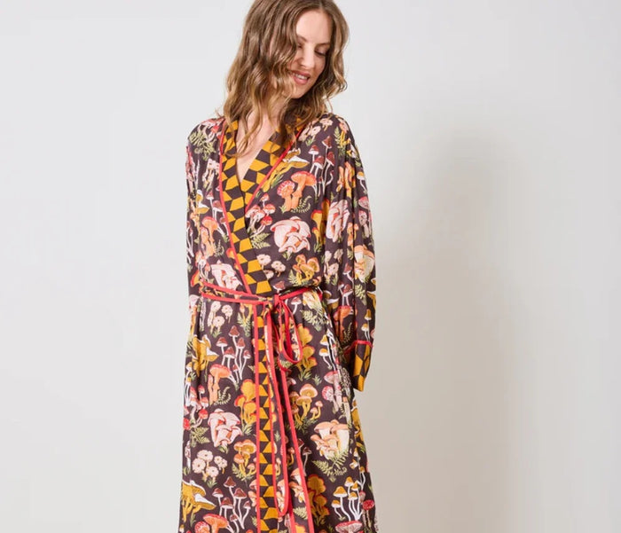 A woman is shown from the waist up wearing the PRINTFRESH - MUSHROOM MEDLEY ECO SATIN ROBE by PRINTFRESH, a kimono-style cottage-core robe featuring yellow geometric and colorful mushroom print patterns, red trim, and a tied belt. She stands against a plain wall.