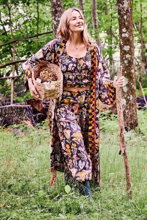 A woman is shown from the waist up wearing the PRINTFRESH - MUSHROOM MEDLEY ECO SATIN ROBE by PRINTFRESH, a kimono-style cottage-core robe featuring yellow geometric and colorful mushroom print patterns, red trim, and a tied belt. She stands against a plain wall.