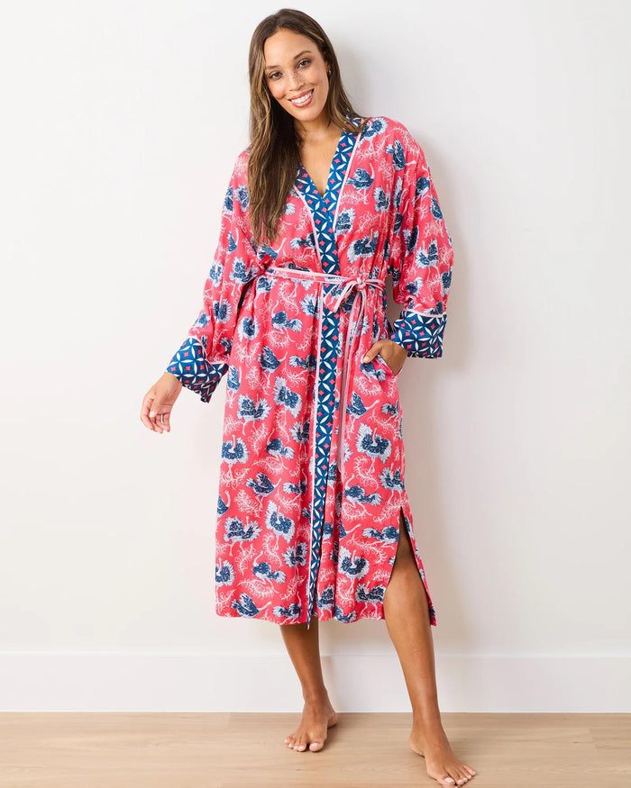 A woman wearing the PRINTFRESH - OSTRICH DANCE SATIN ROBE—an elegant, long pink floral satin robe with blue patterned trim from PRINTFRESH—stands barefoot on a light wood floor against a white wall, smiling at the camera in this sustainable sleepwear look.