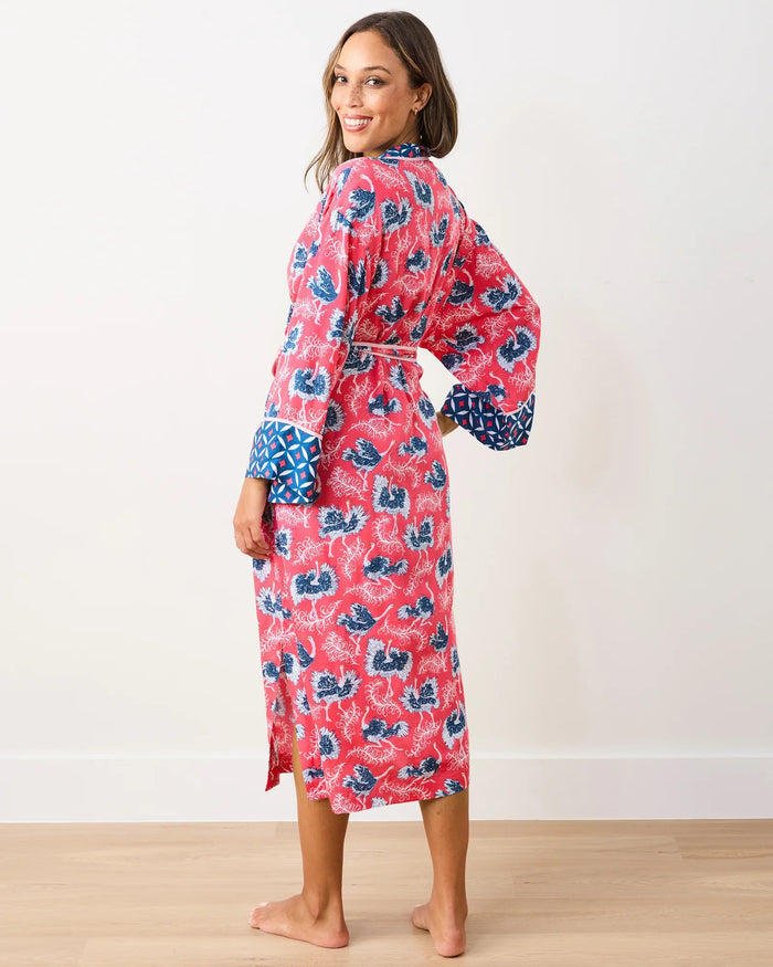 A woman wearing the PRINTFRESH - OSTRICH DANCE SATIN ROBE—an elegant, long pink floral satin robe with blue patterned trim from PRINTFRESH—stands barefoot on a light wood floor against a white wall, smiling at the camera in this sustainable sleepwear look.