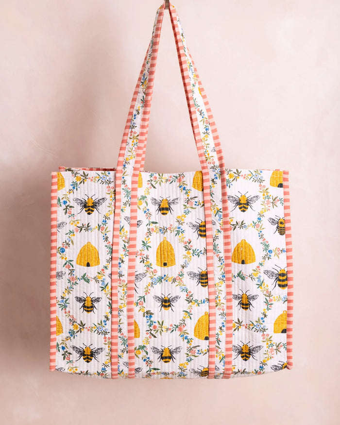 The PRINTFRESH - SWEET BEE TRAVELER TOTE from the Sweet Bee collection showcases floral and bee designs with yellow beehives on white, complemented by pink and white striped handles and borders for a charming contrast against a light beige wall.