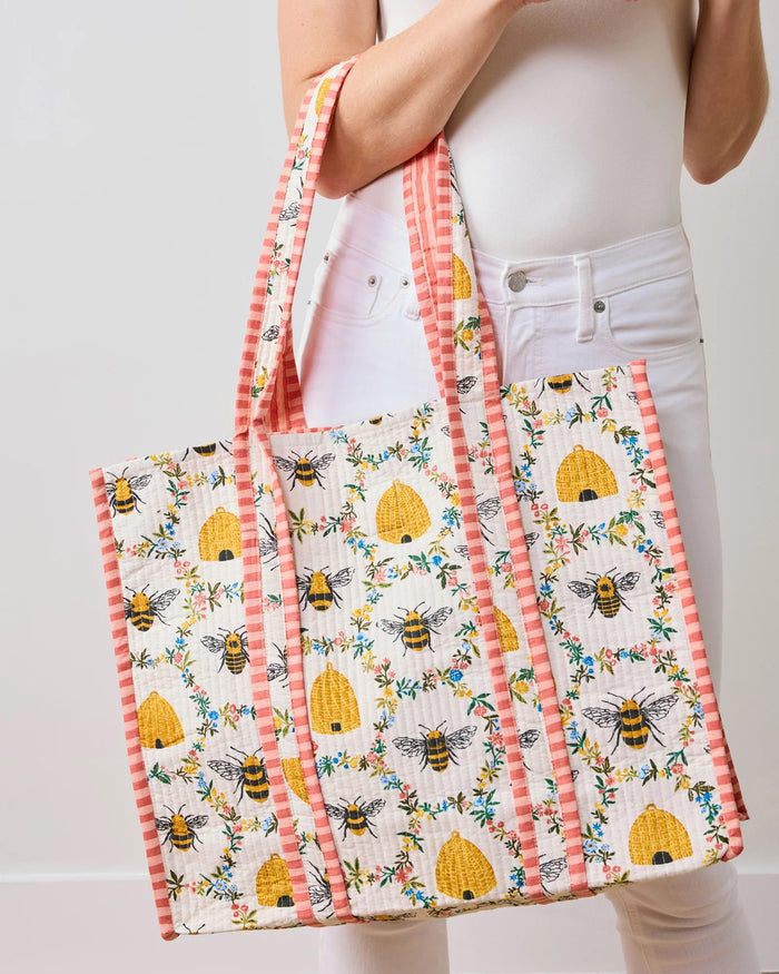 The PRINTFRESH - SWEET BEE TRAVELER TOTE from the Sweet Bee collection showcases floral and bee designs with yellow beehives on white, complemented by pink and white striped handles and borders for a charming contrast against a light beige wall.