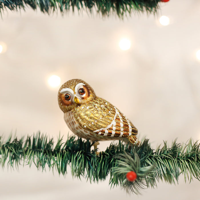 A gold and white owl ornament with glitter sits on a green, festive tree branch with small red berries and blurred lights in the background.