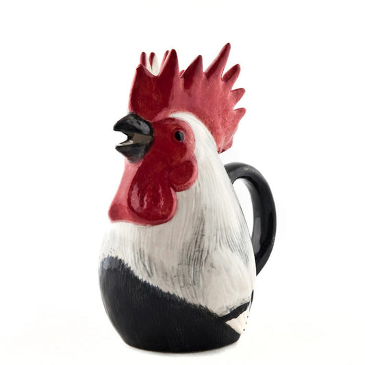 The QUAIL CERAMICS - DORKING ROOSTER JUG by QUAIL DESIGNS, a hand-painted stoneware jug with a red comb and black handle, sits on the table beside a green glass and patterned plates—a charming British tea accessory.