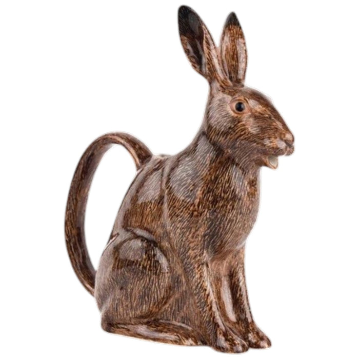 The QUAIL CERAMICS - HARE JUG, LARGE by QUAIL DESIGNS, a delightful ceramic pitcher shaped like a brown rabbit, sits on a wooden surface with white flowers artfully arranged in its open mouth. This charming decorative piece brings whimsy to the tabletop display, while green plants create a softly blurred background.