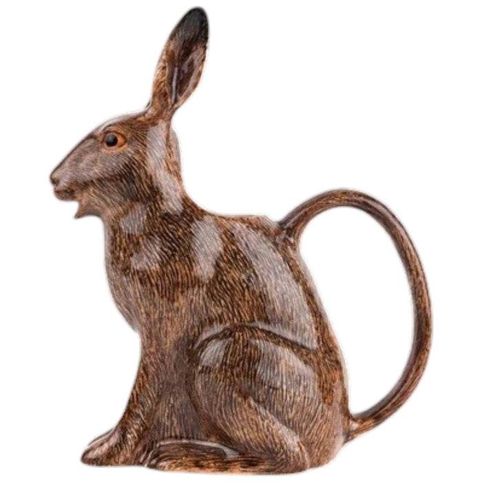 The QUAIL CERAMICS - HARE JUG, LARGE by QUAIL DESIGNS, a delightful ceramic pitcher shaped like a brown rabbit, sits on a wooden surface with white flowers artfully arranged in its open mouth. This charming decorative piece brings whimsy to the tabletop display, while green plants create a softly blurred background.
