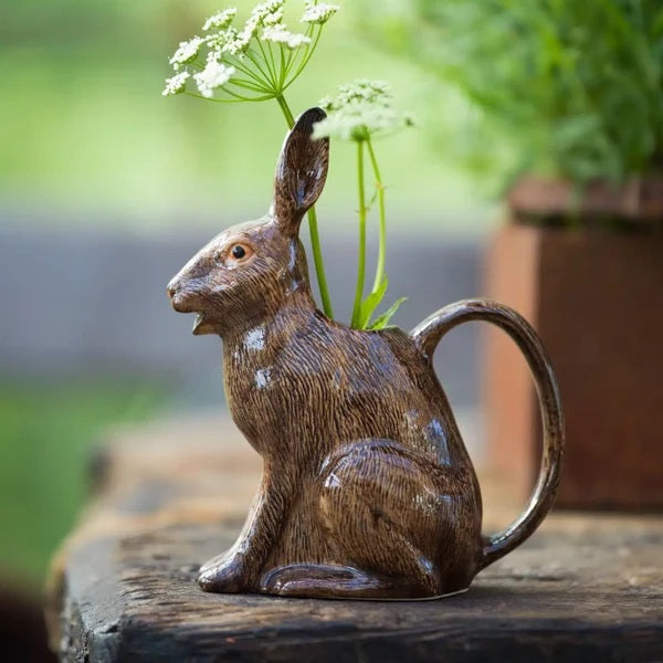 The QUAIL CERAMICS - HARE JUG, LARGE by QUAIL DESIGNS, a delightful ceramic pitcher shaped like a brown rabbit, sits on a wooden surface with white flowers artfully arranged in its open mouth. This charming decorative piece brings whimsy to the tabletop display, while green plants create a softly blurred background.