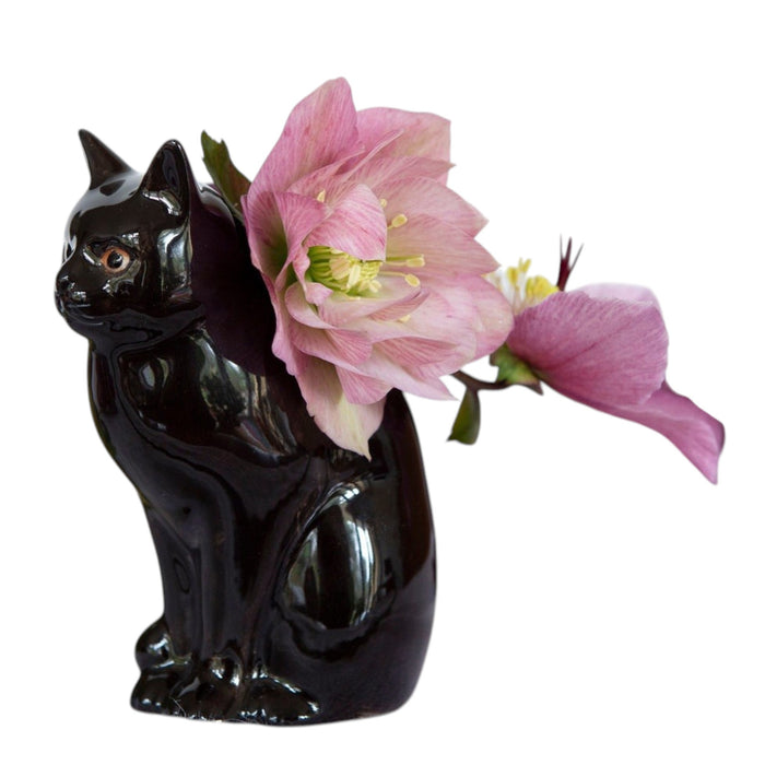 Two QUAIL DESIGNS QUAIL CERAMICS - LUCKY CAT BUD VASES in glossy black stoneware are displayed on a white surface; one holds dark purple and green flowers, while the other sits beside it, both set against a neutral background.