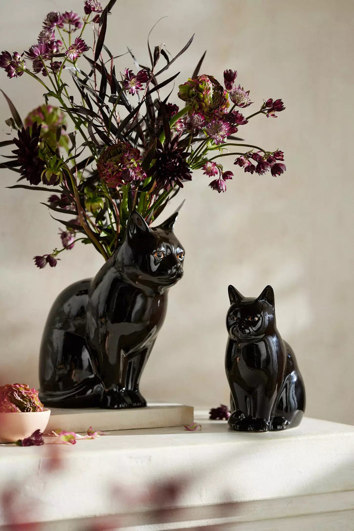 Two QUAIL DESIGNS QUAIL CERAMICS - LUCKY CAT BUD VASES in glossy black stoneware are displayed on a white surface; one holds dark purple and green flowers, while the other sits beside it, both set against a neutral background.