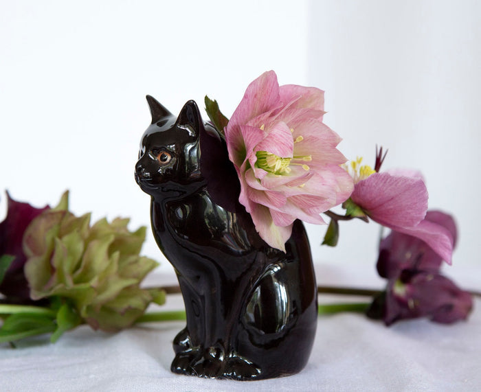 Two QUAIL DESIGNS QUAIL CERAMICS - LUCKY CAT BUD VASES in glossy black stoneware are displayed on a white surface; one holds dark purple and green flowers, while the other sits beside it, both set against a neutral background.
