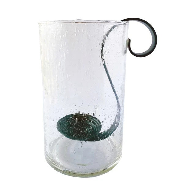 Three upright books are beside the CREATIVE COOP RECYCLED BUBBLE GLASS HURRICANE WITH METAL VOTIVE HOLDER, which contains a lit white candle and small pebbles. The lantern features a black metal handle and sits against a plain light background.
