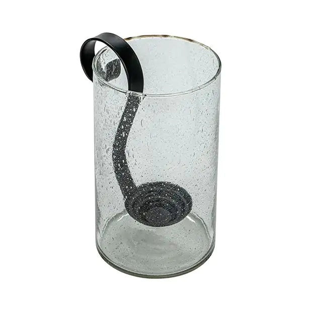 Three upright books are beside the CREATIVE COOP RECYCLED BUBBLE GLASS HURRICANE WITH METAL VOTIVE HOLDER, which contains a lit white candle and small pebbles. The lantern features a black metal handle and sits against a plain light background.
