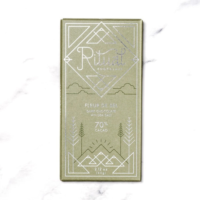 A green RITUAL CHOCOLATE - FLEUR DE SEL CHOCOLATE BAR 70% CACAO with silver geometric designs and text on the wrapper, shown on a white marble background.