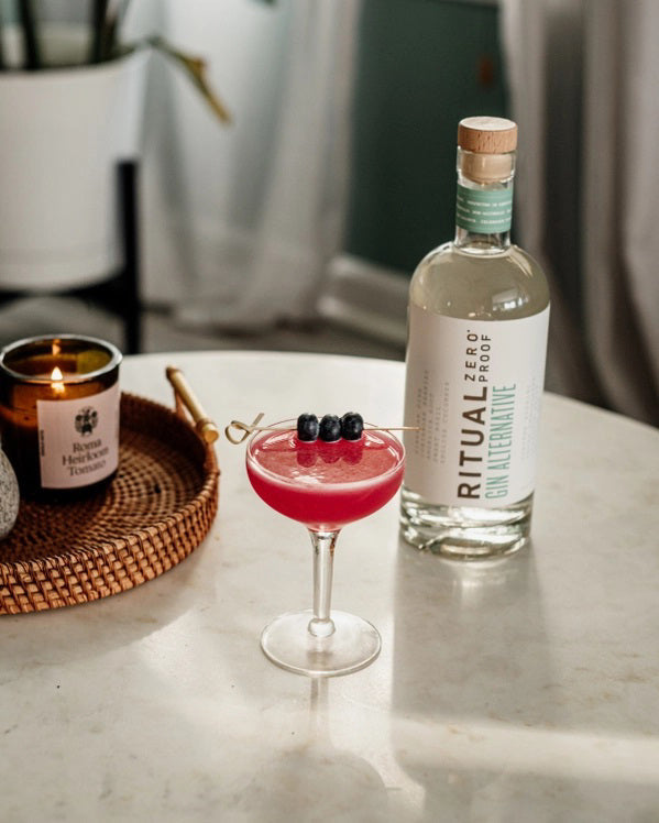 A clear glass bottle of RITUAL ZERO PROOF - GIN ALTERNATIVE from RITUAL ZERO PROOF, featuring a wooden cap and containing a non-alcoholic gin alternative, is displayed against a plain white background.