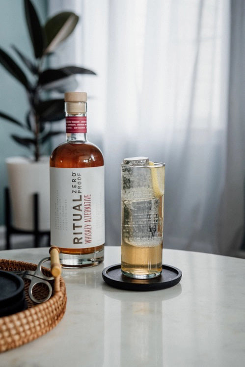 A clear bottle of RITUAL ZERO PROOF - WHISKEY ALTERNATIVE by RITUAL ZERO PROOF, featuring a tan cap and white label, filled with amber liquid and standing upright against a white background.