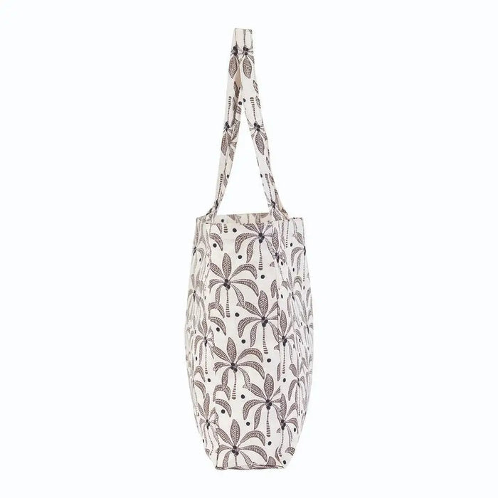 The ROCKFLOWERPAPER PALM SHOPPER TOTE BAG is a spacious cotton canvas bag with two handles, featuring a stylish brown palm tree print on a white background.