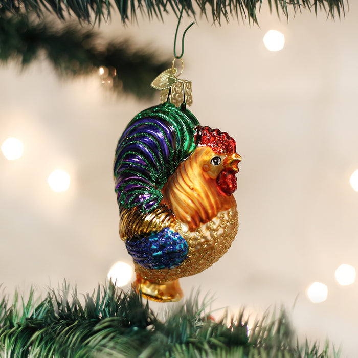 A colorful rooster-shaped Christmas ornament hangs on a green Christmas tree branch with blurred white lights in the background.