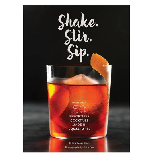 The SHAKE. STIR. SIP. BY KARA NEWMAN recipe book from CHRONICLE BOOKS rests on a light marble surface alongside an orange cocktail with ice, a metal jigger, and a cocktail strainer.