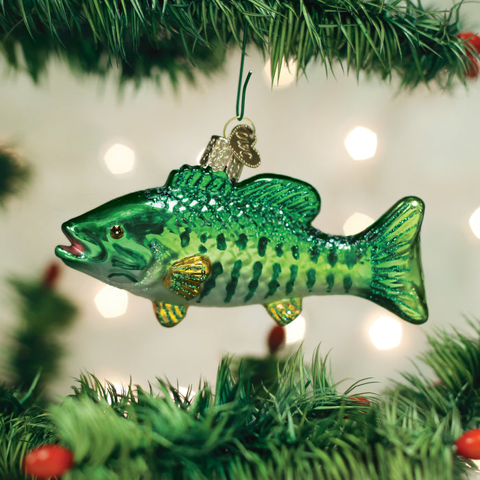 A shiny green fish-shaped ornament hangs from a Christmas tree branch, with blurred white lights in the background.