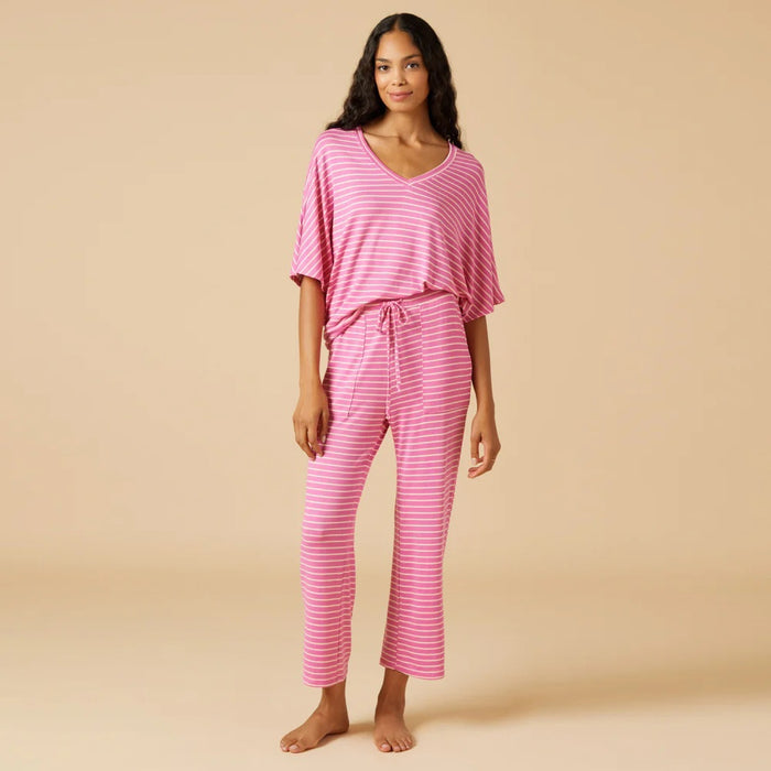 A woman stands barefoot facing away, wearing the SOFTIES Dream Relaxed V-Neck with Capri Lounge Set in Fuschia Stripe—soft jersey pink and white striped pajamas with a loose top and wide-leg pants—against a plain beige background.