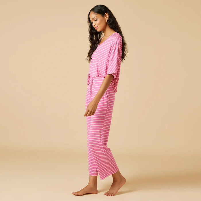 A woman stands barefoot facing away, wearing the SOFTIES Dream Relaxed V-Neck with Capri Lounge Set in Fuschia Stripe—soft jersey pink and white striped pajamas with a loose top and wide-leg pants—against a plain beige background.
