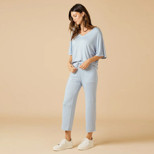 A woman smiles over her shoulder in the SOFTIES DREAM RELAXED V-NECK WITH CAPRI LOUNGE SET IN SKY BLUE STRIPE, paired with white sneakers, against a plain beige background.