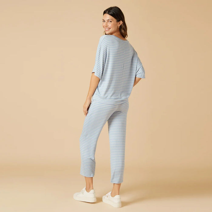 A woman wears the SOFTIES Dream Relaxed V-Neck with Capri Lounge Set in Sky Blue Stripe, featuring soft jersey fabric, short sleeves, matching drawstring capri pants, and white sneakers for a relaxed, all-day comfortable look.