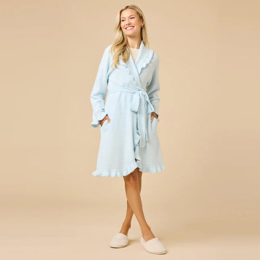A woman stands barefoot on a beige floor, modeling the SOFTIES - RUFFLE CHENILLE ROBE 36" LIGHT BLUE. She faces away with her head turned slightly, hands clasped behind her back, highlighting the robe’s ruffle and belted details.