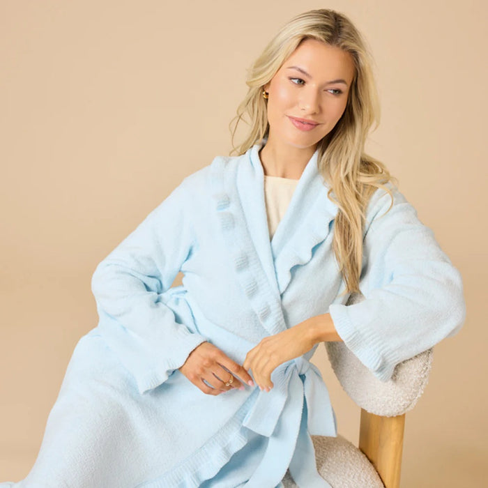 A woman stands barefoot on a beige floor, modeling the SOFTIES - RUFFLE CHENILLE ROBE 36" LIGHT BLUE. She faces away with her head turned slightly, hands clasped behind her back, highlighting the robe’s ruffle and belted details.