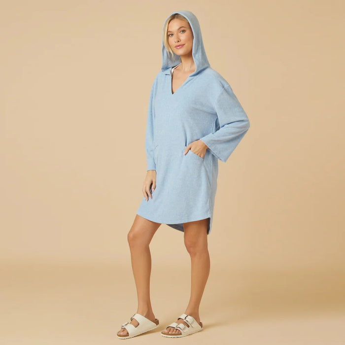 A woman faces away in the SOFTIES Terry Hooded Lounger in Heather Sky Blue, paired with white sandals. Her blonde ponytail grazes a visible bikini top, blending resort-ready comfort and style against a beige backdrop.