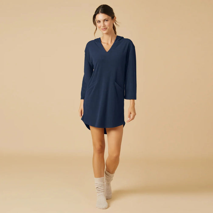 A woman stands on a beige background wearing the SOFTIES Terry Hooded Lounger in Midnight, paired with light gray socks. With her hair tied back and a slight smile, she showcases the brand's signature resort-ready comfort in this stylish look.