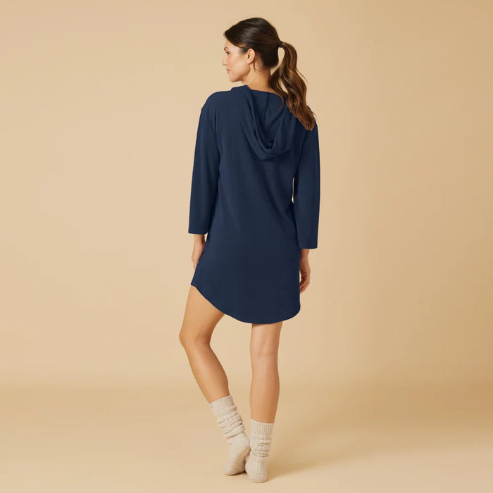 A woman stands on a beige background wearing the SOFTIES Terry Hooded Lounger in Midnight, paired with light gray socks. With her hair tied back and a slight smile, she showcases the brand's signature resort-ready comfort in this stylish look.