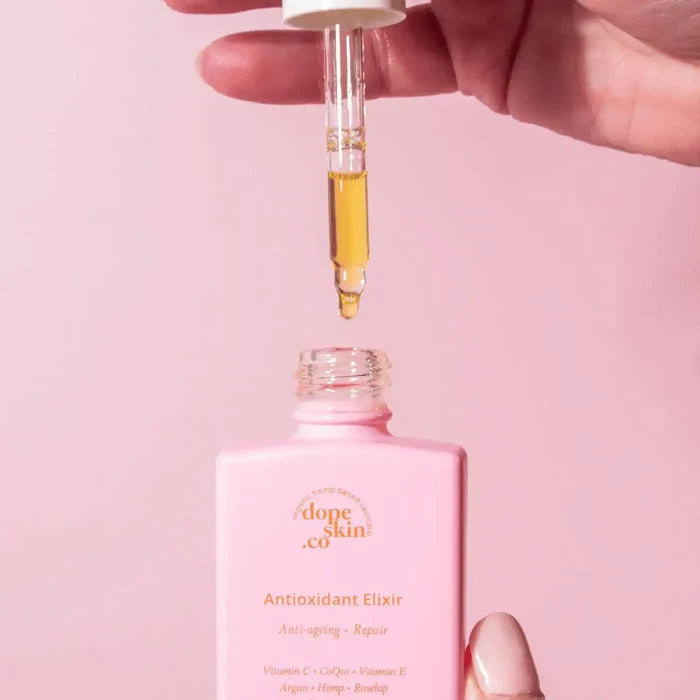 Hand dropping a dropper of yellow liquid into a pink bottle labeled 'dope skin co. Antioxidant Elixir' on a pink background.