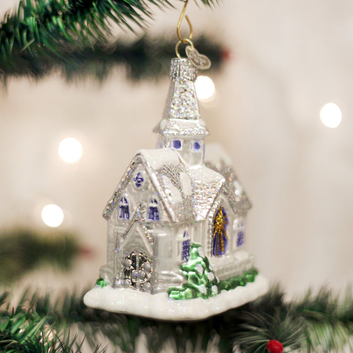 A sparkling church-shaped Christmas ornament with snowy details hangs from a green tree branch, surrounded by soft, glowing white lights in the background.