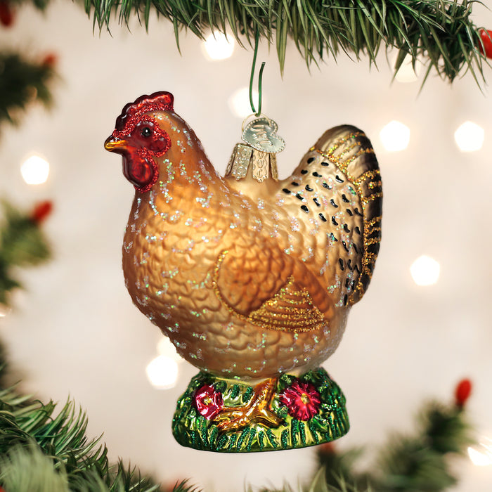 A glass ornament shaped like a brown chicken with gold and black detailing hangs from a Christmas tree branch, surrounded by festive lights.