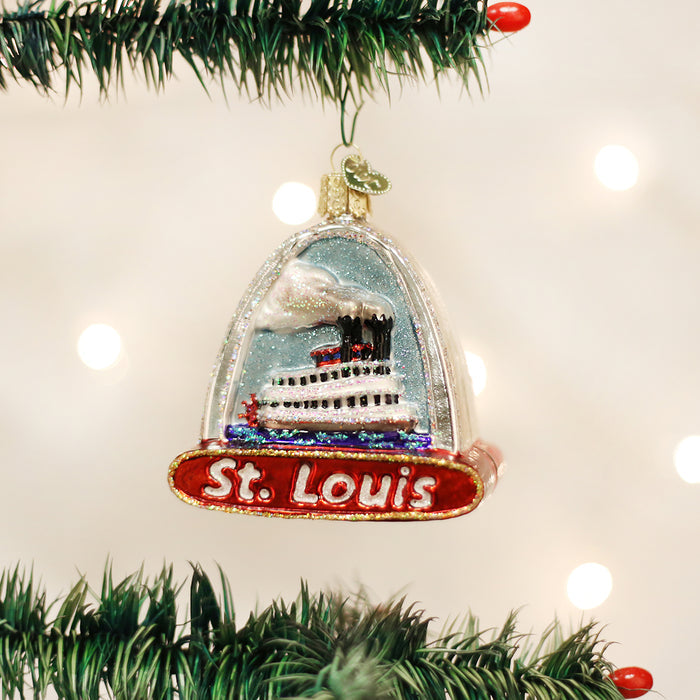 A festive Christmas ornament shaped like a riverboat sits inside a clear arch labeled St. Louis, hanging on a green tree with blurred white lights in the background.