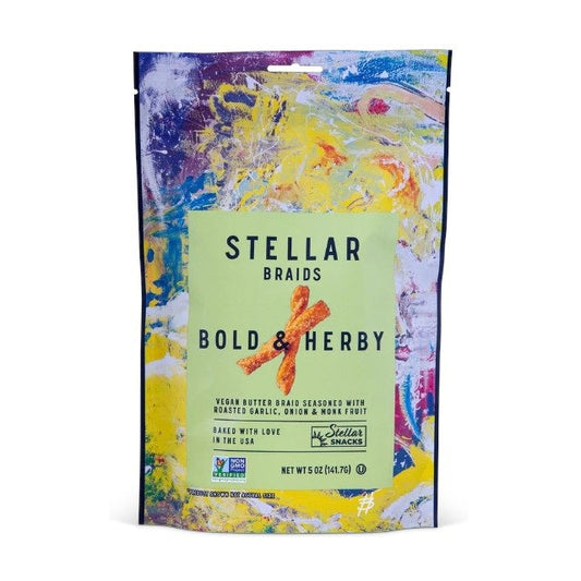 A bowl of STELLAR SNACKS - BOLD AND HERBY PRETZEL BRAIDS rests on a wooden board beside a yellow bag labeled with the STELLAR SNACKS brand name. Some pretzel braids, featuring their roasted garlic and onion flavor, are scattered on the board, while a beige cloth is partially visible.