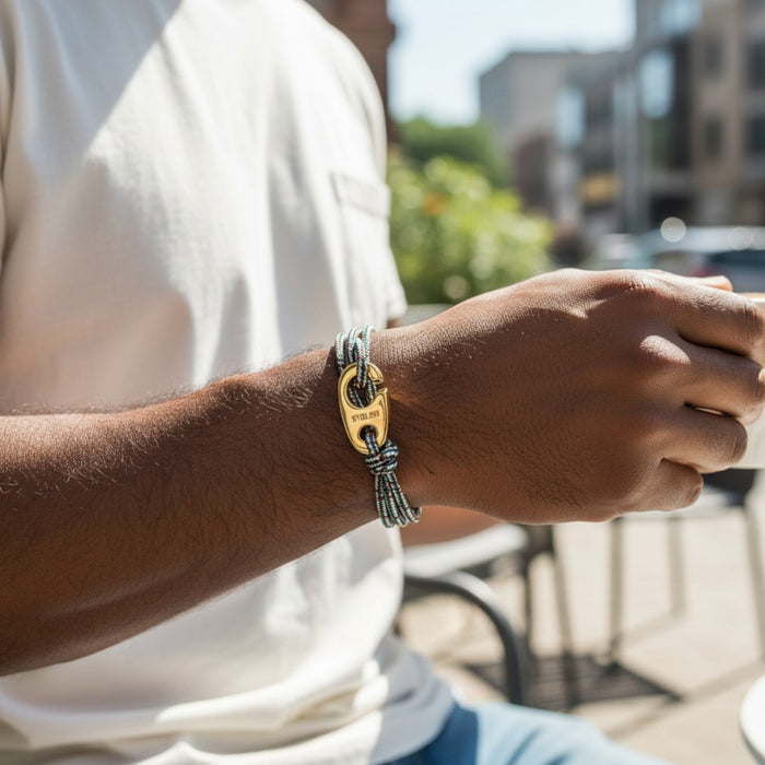 Sitting outdoors in sunlight, a person in a white shirt holds a cup while wearing the STOLEN RICHES BYRON Rope Bracelet with Gold Brummel Hook—a stylish, waterproof accessory. Blurred urban scenery forms the backdrop.