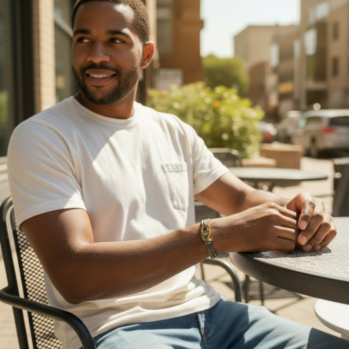 Sitting outdoors in sunlight, a person in a white shirt holds a cup while wearing the STOLEN RICHES BYRON Rope Bracelet with Gold Brummel Hook—a stylish, waterproof accessory. Blurred urban scenery forms the backdrop.