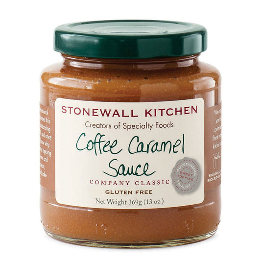 A jar of STONEWALL KITCHEN - COFFEE CARAMEL SAUCE is displayed alongside a stick of butter, brown sugar, coffee beans, a glass of syrup, a small glass of oil, and a metal cup of cream on a wooden surface. This decadent caramel sauce from STONEWALL KITCHEN provides a rich and buttery sweetness that’s perfect for elevating your favorite desserts.